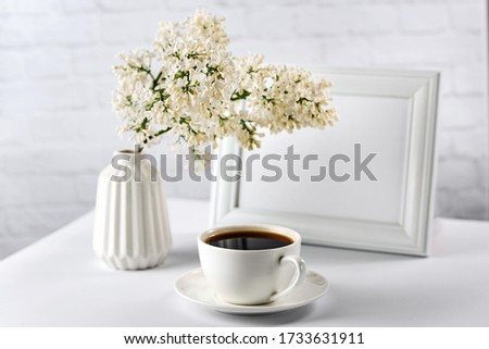 Still life with white objects on a white background. Blank photo frame, vase with flowers and a cup of coffee. Layout for the morning concept.