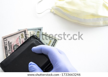 Close up of person holding a black  leather wallet wearing a blue latex glove with a twenty, fifty and hundred dollar bill sticking out and fanned with yellow mask on white background with copy space.