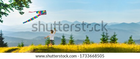 International Children's Day concept. Little happy boy running with a colored striped kite on a background of beautiful mountain landscape. Active outdoor entertainment, healthy children.