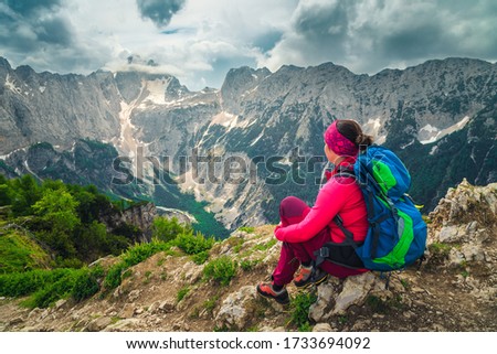 Cheerful hiker woman with colorful backpack. Freedom hiker woman sitting and enjoying the view with Jalovec mountain peak, from the Slemenova Spica hiking place, Julian Alps, Slovenia, Europe Royalty-Free Stock Photo #1733694092