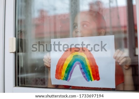 Little girl in the window holds a picture of a rainbow symbolizing the imminent end of the quarantine in connection with the coronavirus pandemic.