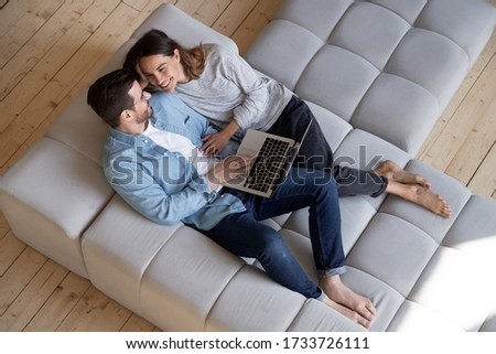 Top view of happy millennial couple lying relaxing on modern design comfortable couch in new home, smiling young husband and wife rest on cozy sofa using laptop enjoy family weekend together