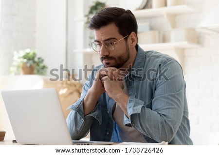 Pensive young Caucasian man in glasses sit at desk look at laptop screen thinking pondering, thoughtful millennial male in spectacles work at computer consider ideas solving problems at home workplace Royalty-Free Stock Photo #1733726426