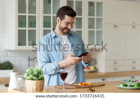 Smiling Caucasian young man use cellphone gadget texting messaging while preparing food in kitchen, happy millennial male read recipe on smartphone get ready for home party or celebration