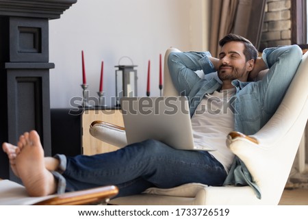 Happy millennial man sit relax in comfortable design armchair in living room take nap distracted from computer work, smiling young male rest in chair at home breathe fresh air, stress free concept Royalty-Free Stock Photo #1733726519
