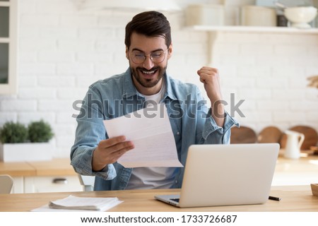 Excited millennial man in glasses sit at table in home kitchen feel euphoric reading good news in postal letter, overjoyed young male triumph receive great unexpected answer in post correspondence Royalty-Free Stock Photo #1733726687