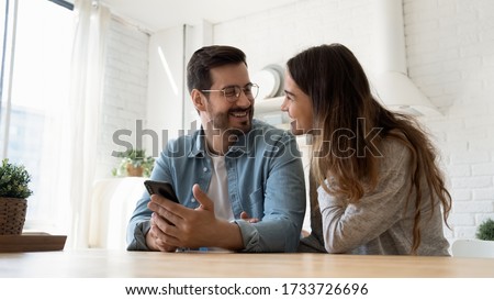 Overjoyed husband and wife sit at table in modern design kitchen laugh enjoy family weekend using modern smartphone gadget together, happy couple relax at home have fun browsing Internet on cell