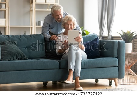 Full length happy mature grey-haired woman sitting on couch, showing wished product in internet store on digital computer tablet to smiling elderly retired husband, shopping together online at home.