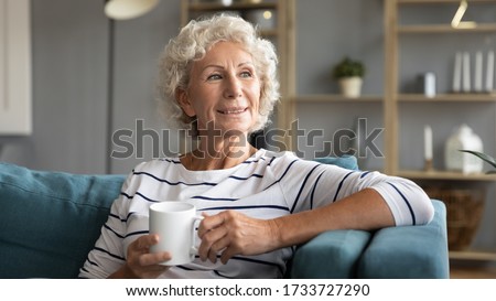Head shot smiling old senior woman relaxing on comfortable couch with mug of hot tea, enjoying free leisure weekend time alone in living room. Happy mature grandmother drinking morning coffee.
