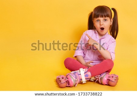 Picture of astonished female child with widely opened mouth sitting on floor with crossed legs, pointing with her forefinger aside, showing something amazing. Copy space for advertisement or promotion