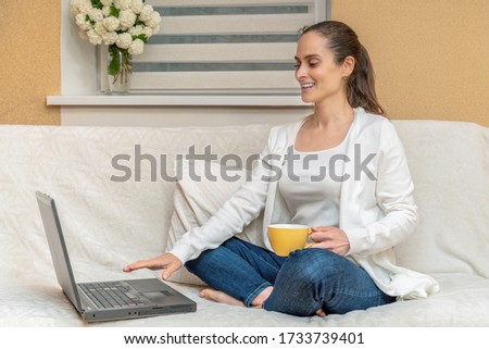 A woman in jeans and a white jacket on the sofa drinking a Cup of hot coffee or tea using a laptop a woman checks social apps and works. Communication and technology concept