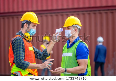 Factory worker man checking fever by digital thermometer for scan and protect from Coronavirus (COVID-19) at cargo containers - Healthcare Concept Royalty-Free Stock Photo #1733743910