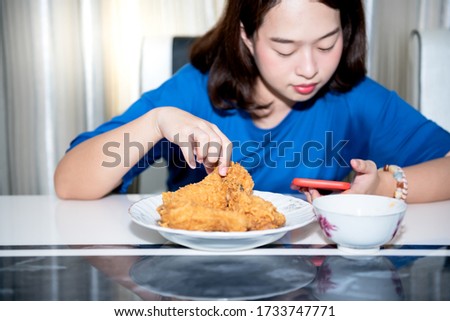 A 22 year old Asian woman is using her hand to pick up fried chicken placed on a plate, While she is interested in a mobile phone, This picture focused on the hand. to food concept.