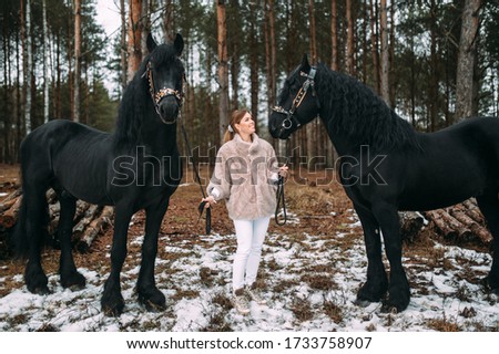 Magnetic young female in grey fur coat and white trousers stands with two big black horses outside.