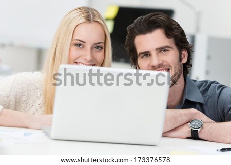 Young couple working on a laptop at home crouching down in the living room smiling at the camera over the top of the screen