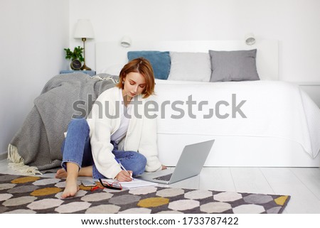 Woman sitting on floor indoors, near laptop computer, writing, taking notes.  Remote working, creating, relax and leisure. Online studying or freelance working at home in cozy interior apartment Royalty-Free Stock Photo #1733787422