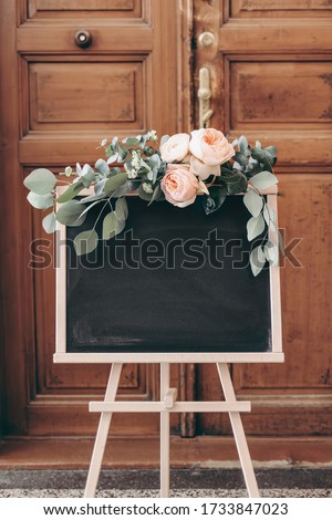 Blank wedding chalkboard sign mockup scene. Wooden easel with welcome board. Floral garland of green eucalyptus branches and English roses flowers. Rustic birthday party decoration. Blurred old door.