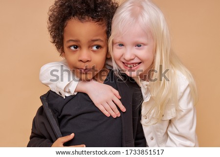 two friendly multiracial children stand together isolated over beige background, diverse positive boy and girl stand hugging. children concept