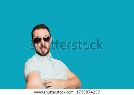 A young bearded man is fooling around at the camera on a blue background. The guy takes a selfie and builds funny faces
