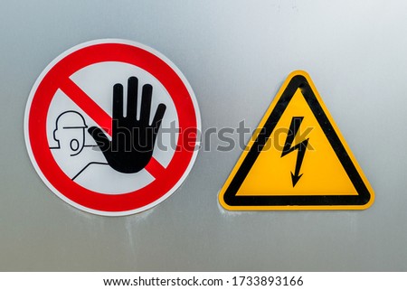 On a giant iron door two warning signs are placed: a round red and white prohibition sign with a don't touch symbol and a yellow triangular shaped sign with a high voltage symbol