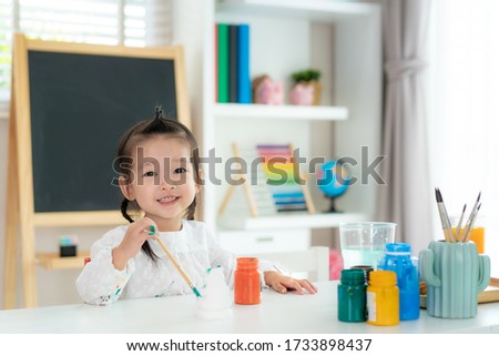 Asian kindergarten school girl looking at camera and smile while painting Plaster doll with Acrylic water color paint in living room at home. Homeschooling and distance learning.
