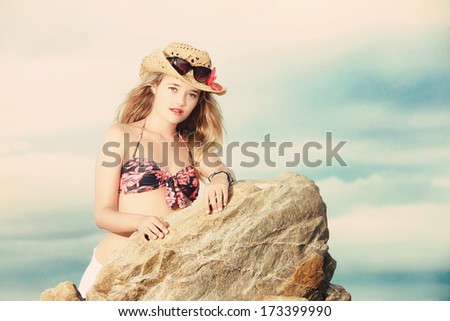 A closeup photo of a beautiful blonde lady dressed in a floral or tropical bikini with cowboy hat and sunglasses leaning sideways on a rock.