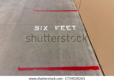 Looking down on sign stenciled in white capital letters stating SIX FEET to indicate social distancing guidelines. Red line is visable as social distance marking