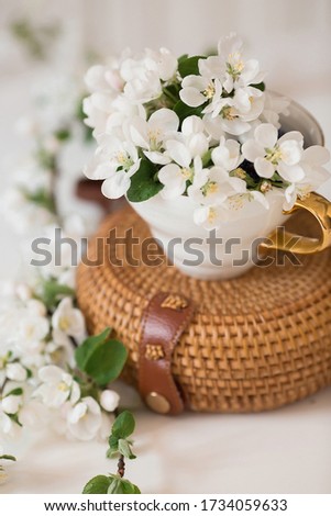 
Delicate, white flowers of an apple tree in a porcelain cup with a golden handle. The dishes are on a textured rattan bag. The concept of spring, ecology and good morning. Copy space for text.