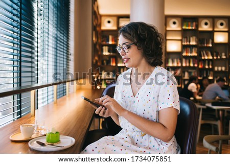 Young female in eyeglasses and light dress surfing mobile phone while sitting with dessert and cup of hot drink in cafe