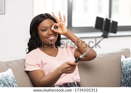 technology and people concept - happy smiling african american woman with smartphone and selfie stick taking picture and looking through ok hand sign at home