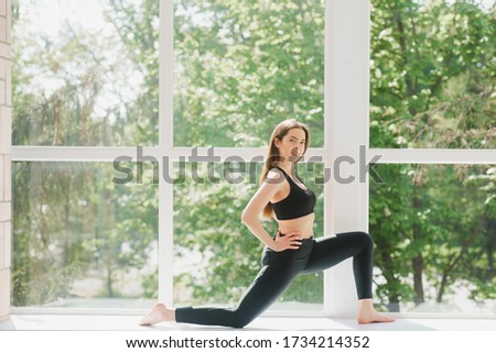 Beautiful woman in grey sportswear, bra and leggings practicing yoga, standing in anjaneyasana pose, girl doing Horse rider exercise, working out at home or in yoga studio with white walls
