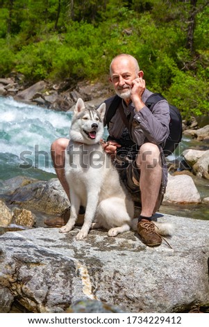 An old man and sled dog walk near the river. Alpine landscape. Active leisure pensioner. An elderly man is smiling. Walk with Siberian Husky.