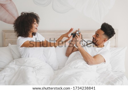 Young african loving couple girl guy in white t-shirts lying on bed doing shot on retro vintage photo camera in bedroom at home, spend time in room. Rest relax good mood quarantine lifestyle concept