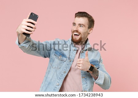 Cheerful young bearded man guy in denim casual jacket posing isolated on pastel pink wall background. People lifestyle concept. Mock up copy space. Doing selfie shot on mobile phone, showing thumb up