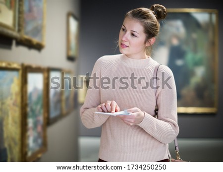 Young pensive woman visitor with guide book looking at exhibition in art museum