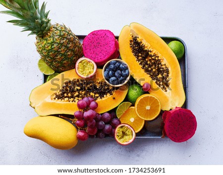 Tropical fruits flat lay with mango, papaya, pitahaya, passion fruit, grapes, limes and pineapples on a tray. Table with ingredients for summer snacks on concrete background. Organic healthy plate