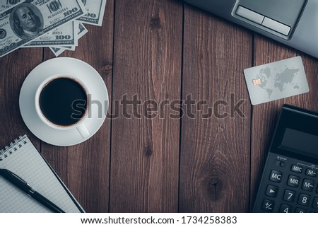 Top view on a wooden rustic table with a cup of coffee, a credit card, a calculator, a notebook with a pen, dollar bills and a laptop with copy space.