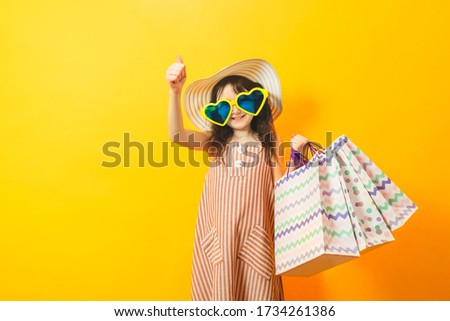 Kid girl playing shopping in the store. Child holding colored paper shopping bags. Big glasses in the shape of hearts. Best buy concept. Yellow studio bright background.