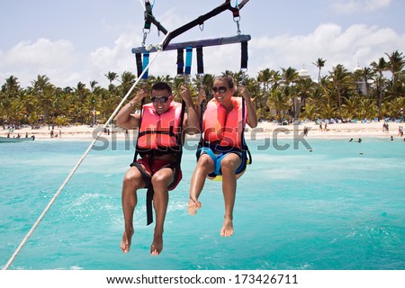 Happy couple Parasailing in Dominicana beach in summer. Couple under parachute hanging mid air. Having fun. Tropical Paradise.