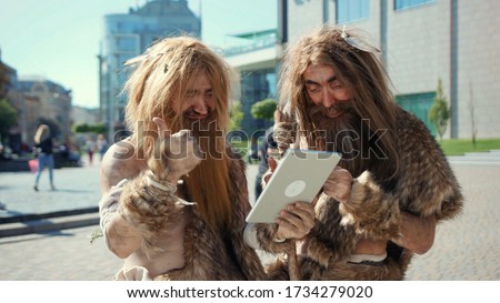 Excited prehistoric bushmen of hunter-gatherers covered in fur browsing internet on tablet discovering technology in modern city. Adaptation concept. Royalty-Free Stock Photo #1734279020