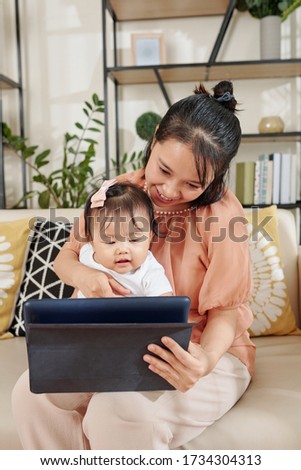Happy young Vietnamese woman showing educational video or application on tablet computer to baby girl