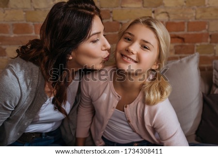 Mother kissing smiling kid looking at camera on couch