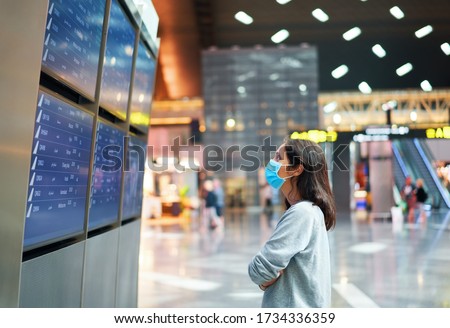 Woman in virus protection face mask looking at information board checking her flight in international airport. Departure board, flight status                              Royalty-Free Stock Photo #1734336359