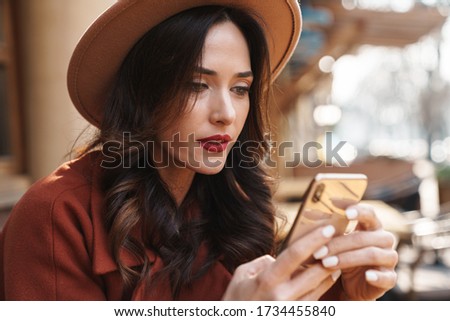 Image of focused elegant adult woman in hat using mobile phone while sitting at street cafe outdoors