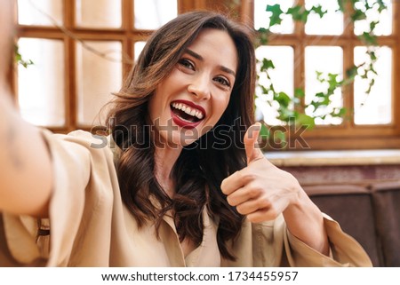 Image of joyful adult woman showing thumb up and taking selfie photo while sitting in cozy cafe indoors