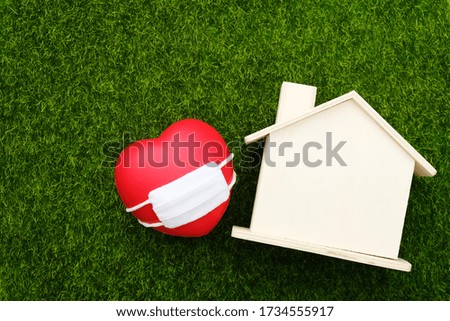Red heart wearing white mask and wooden house model on green grass background. Concept for healthcare insurance, coronavirus self quarantine, or staying at home and social distancing
