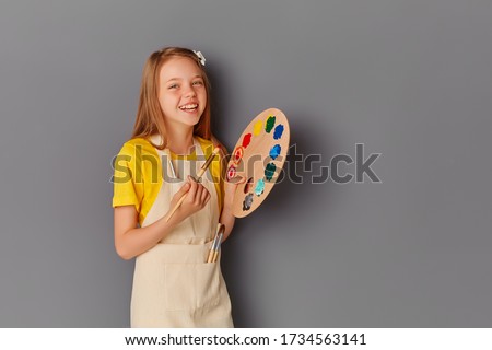 Portrait of a beautiful young teenage girl holding a wooden palette and brush against a gray wall. A smiling child with an art palette and an artist's brush in his hands. Items for children's Royalty-Free Stock Photo #1734563141