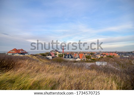 A small town on the Dutch sea coast. In the background a red lighthouse. The houses have gable roofs and nestle into the dunes. In the foreground dunes covered with grass.