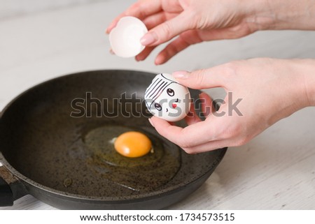 Female hands hold broken white eggshell, cruel murder of egg with drawn face concept, make fried breakfast meal on pan