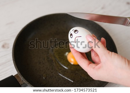 Woman hand with knife going to broke eggshell with drawn by marker upset face on egg, prepare simple protein fried breakfast at pan